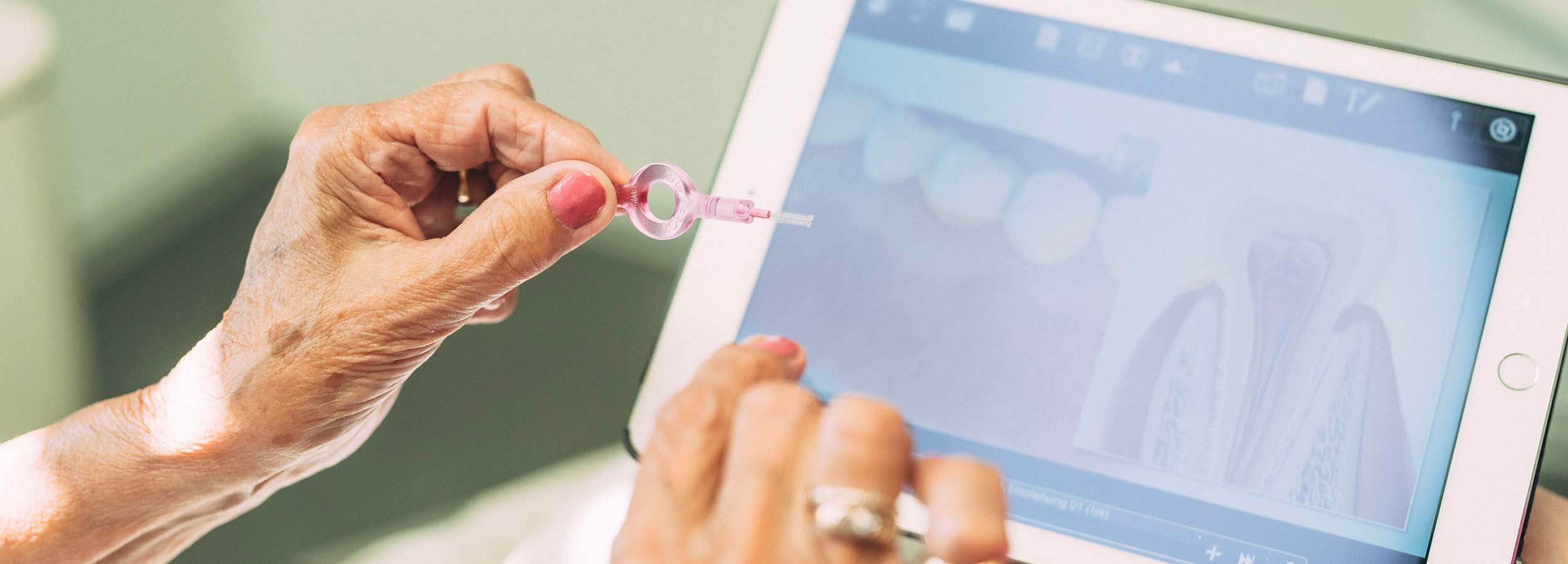 Bildschirm eines Tablets und eine Hand mit Zahnzwischenraumbürste (Interdentalbürste): Auf dem Bildschirm erscheinen stilisierte Zähne, hier wird erklärt, wie richtige Zahnpflege Parodontitis vorbeugen kann.